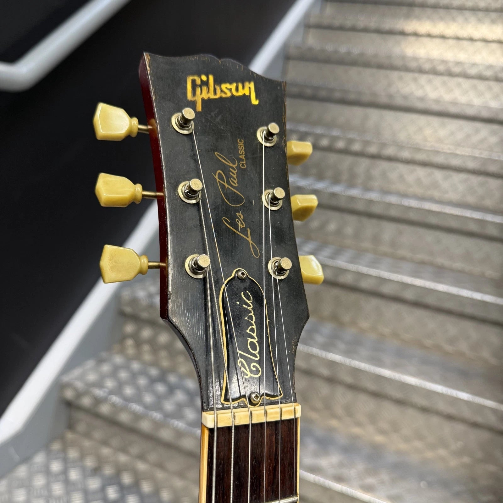 Pre-Owned Gibson Les Paul Classic '1960' 2001 Electric - Cherry Sunburst Close-up of a Gibson Lues Paul Classic acoustic guitar headstock, showing the gold Gibson logo, model script, and six tuning pegs against a blurred staircase background.