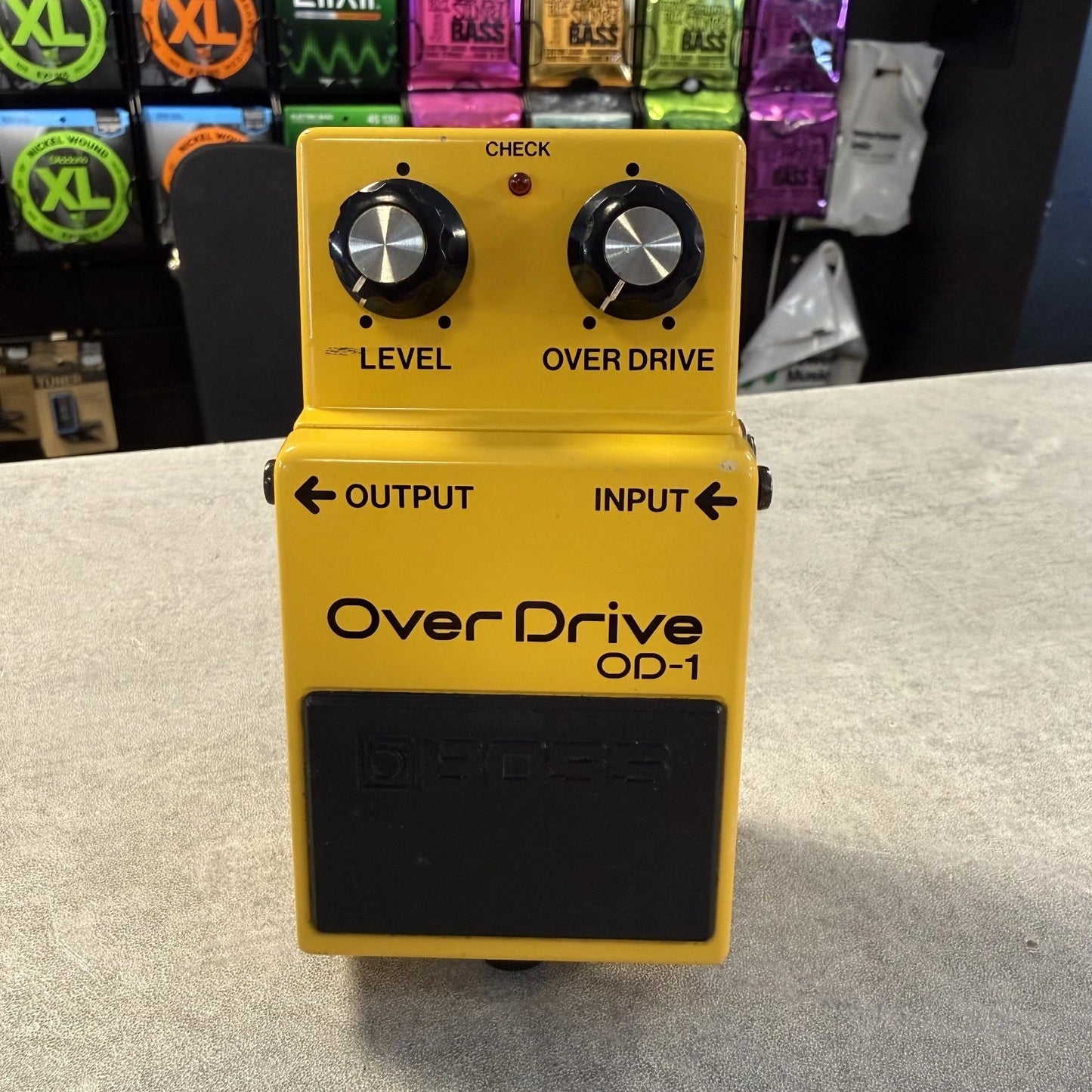 Pre-Owned Boss OD-1 Yellow OverDrive OD-1 guitar effects pedal on a gray surface with a store shelf in the background.