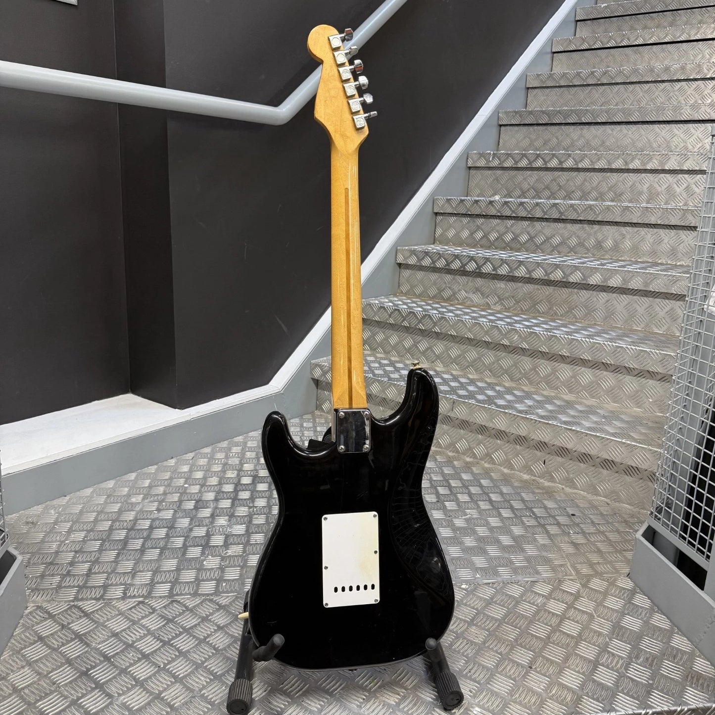 Pre-Owned Fender USA American Traditional Fat Stratocaster (Export Model) 1999 Electric - Black A black electric guitar with a natural wood neck stands on a stand in front of a metal staircase.