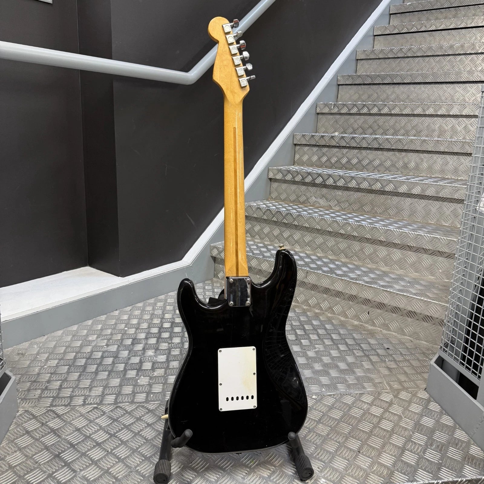 Pre-Owned Fender USA American Traditional Fat Stratocaster (Export Model) 1999 Electric - Black A black electric guitar with a natural wood neck stands on a stand in front of a metal staircase.