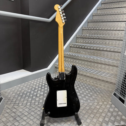 Pre-Owned Fender USA American Traditional Fat Stratocaster (Export Model) 1999 Electric - Black A black electric guitar with a natural wood neck stands on a stand in front of a metal staircase.