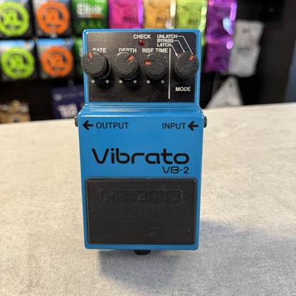 Pre-Owned Boss VB-2 Blue Boss Vibrato VB-2 guitar effects pedal on a gray surface with colorful boxes in the background.
