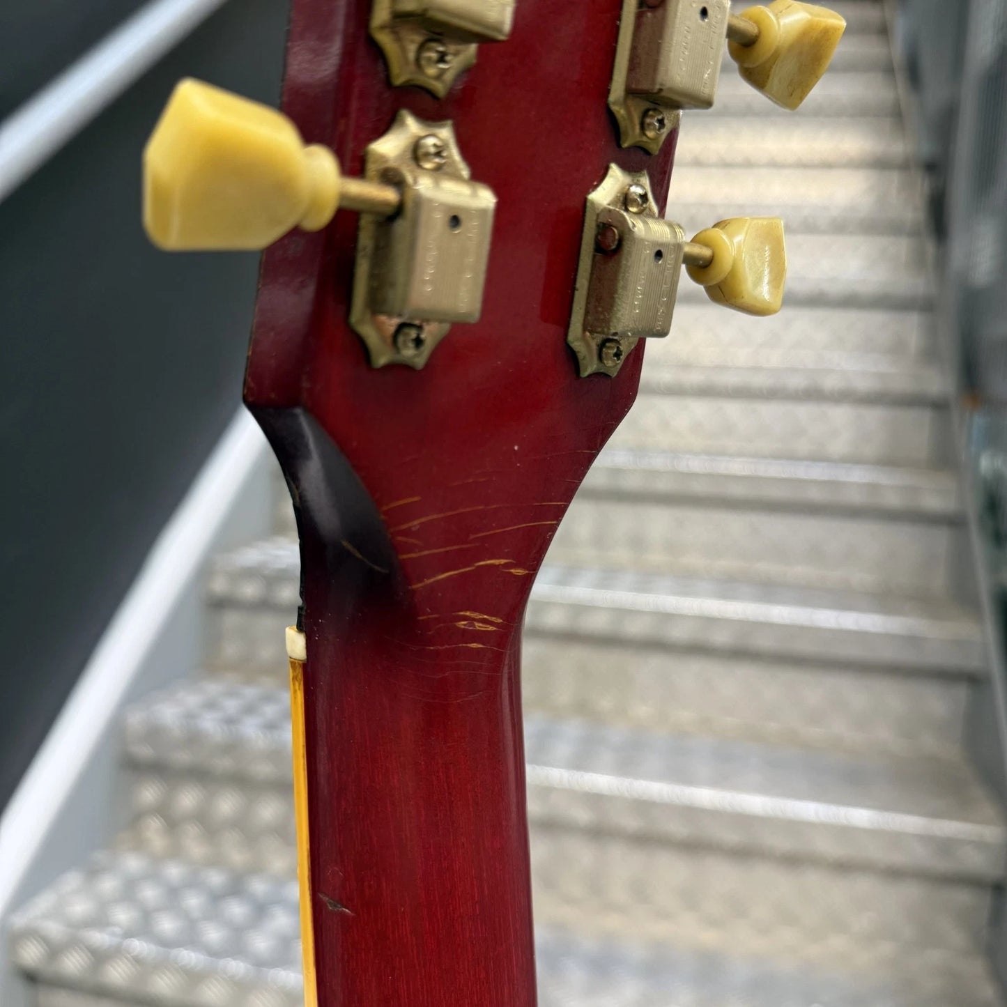 Pre-Owned Gibson Les Paul Classic '1960' 2001 Electric - Cherry Sunburst Close-up of the headstock of a red electric guitar, showing four gold-toned tuning pegs and hardware, with a blurred staircase in the background.