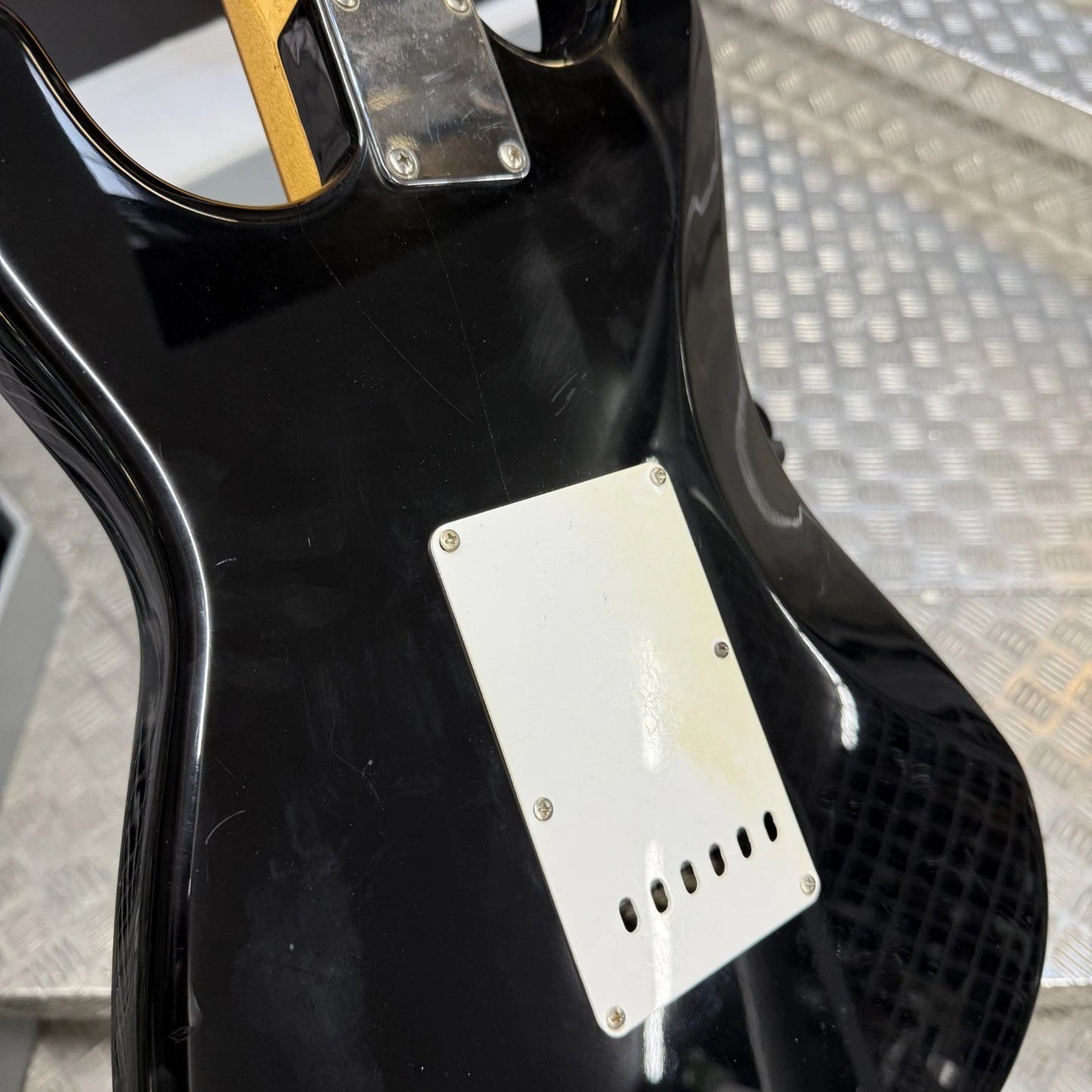 Pre-Owned Fender USA American Traditional Fat Stratocaster (Export Model) 1999 Electric - Black Close-up of the back of a glossy black electric guitar body with a white pickguard and metal hardware, photographed on a textured metal surface.
