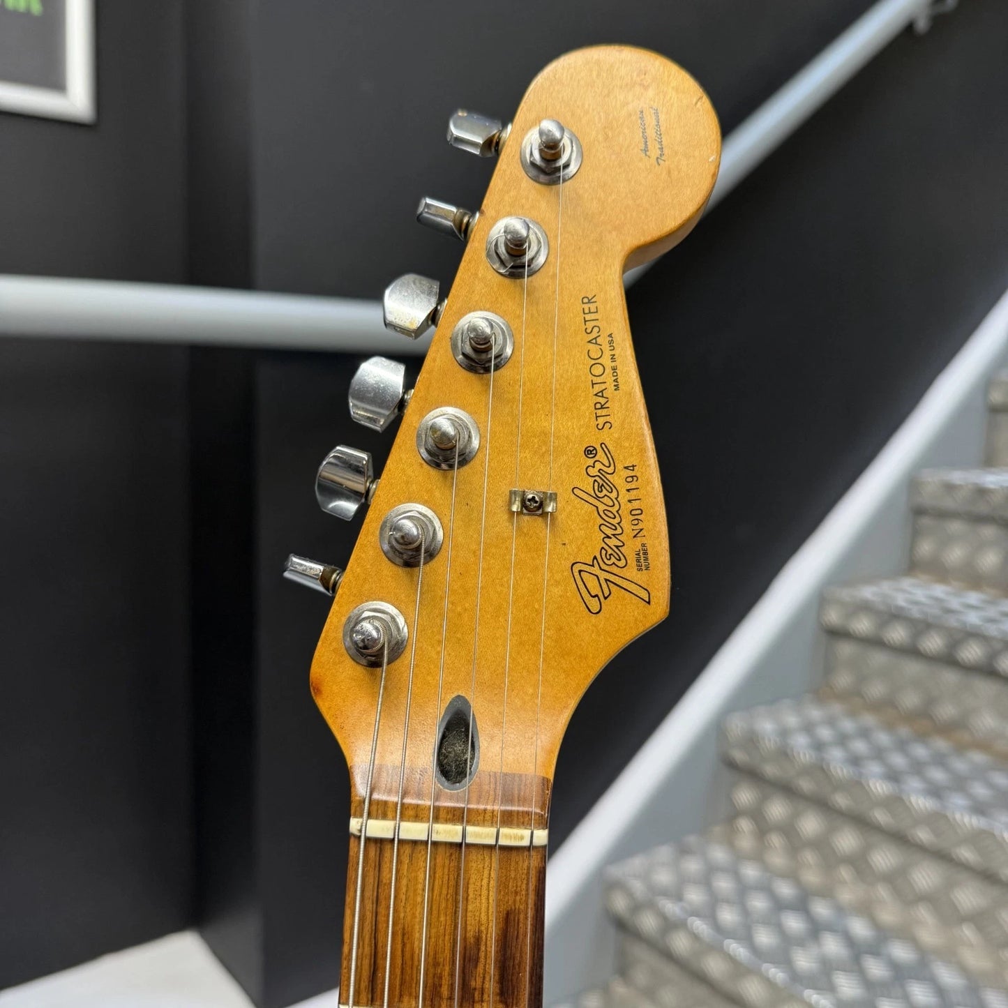 Pre-Owned Fender USA American Traditional Fat Stratocaster (Export Model) 1999 Electric - Black Close-up of a Fender Stratocaster guitar headstock in a natural wood finish, showing six silver tuning pegs and the Fender logo, with a staircase and dark wall in the background.