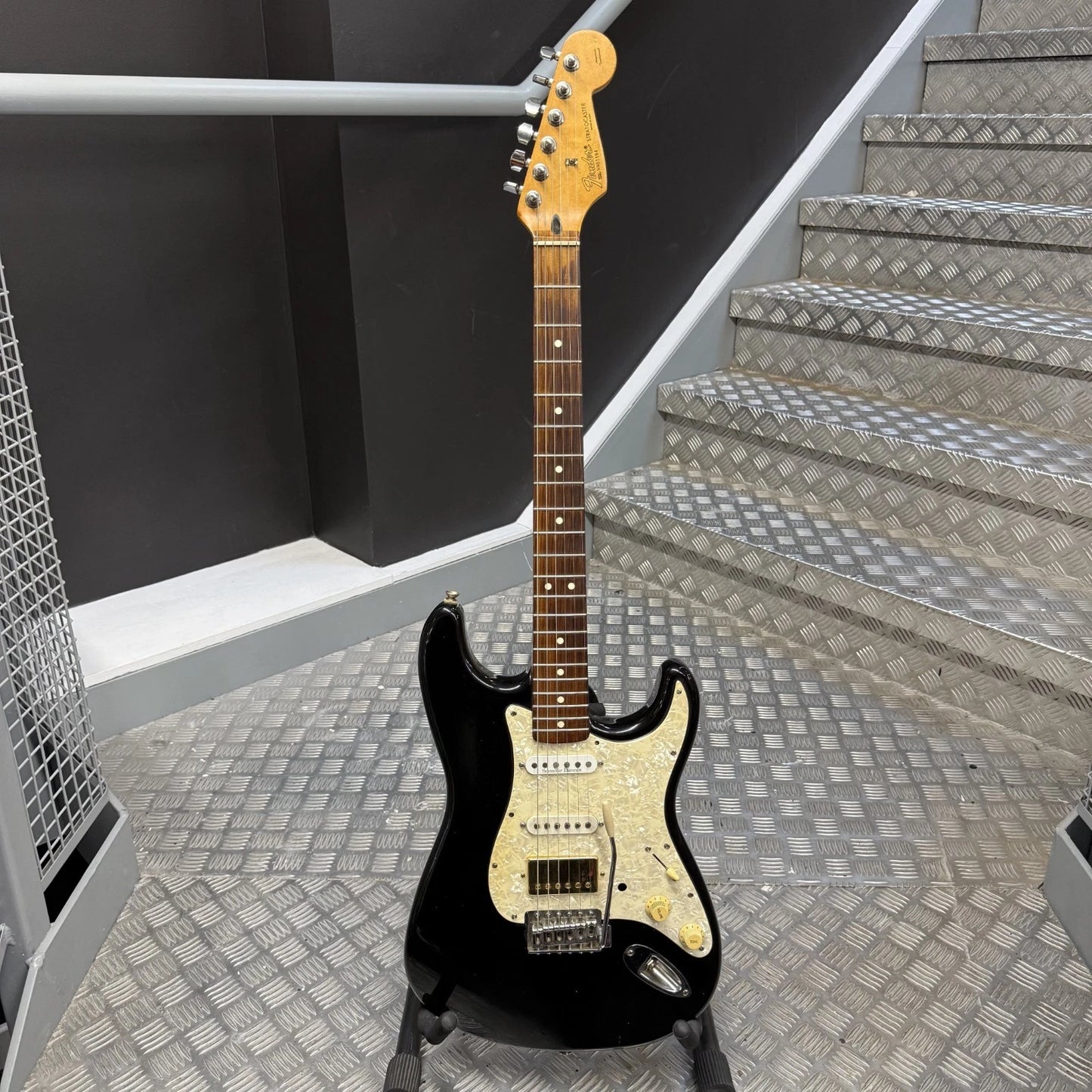 Pre-Owned Fender USA American Traditional Fat Stratocaster (Export Model) 1999 Electric - Black A black electric guitar with a cream pickguard and maple headstock, standing on a stand in front of a staircase with diamond-plate metal steps.