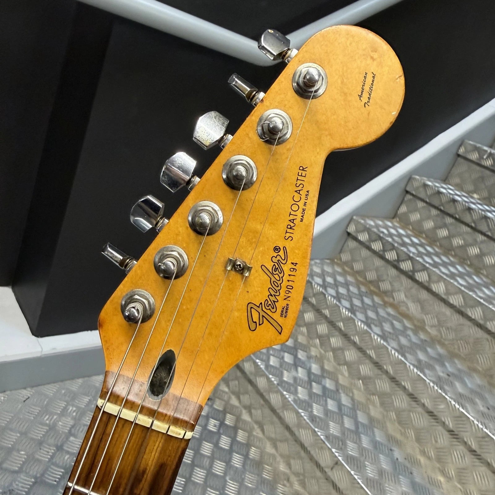 Pre-Owned Fender USA American Traditional Fat Stratocaster (Export Model) 1999 Electric - Black Close-up of a Fender Stratocaster guitar headstock with six silver tuning pegs, showing the gold-toned maple wood headstock and dark brown fretboard against a background of black and gray metal steps.