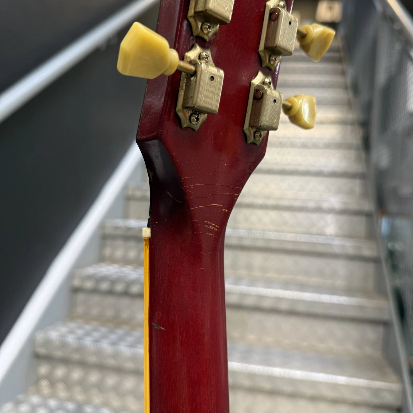 Pre-Owned Gibson Les Paul Classic '1960' 2001 Electric - Cherry Sunburst Close-up of the headstock of a guitar with a dark red finish and cream-colored tuning pegs, set against a blurred staircase background.