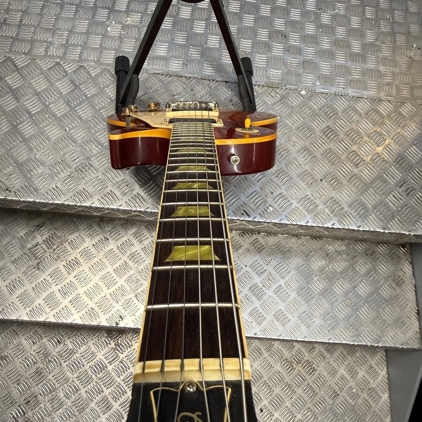 Pre-Owned Gibson Les Paul Classic '1960' 2001 Electric - Cherry Sunburst A close-up of an electric guitar neck and headstock hanging against a diamond plate metal background. The guitar has a dark reddish-brown body with gold binding, a dark fretboard with gold block inlays, and a black pickguard.