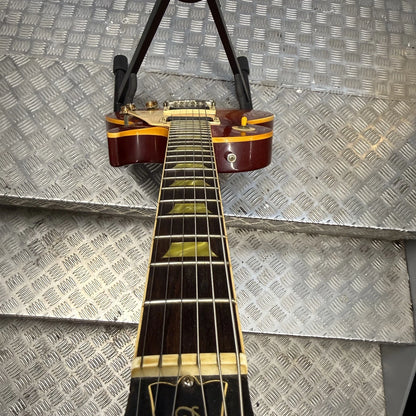Pre-Owned Gibson Les Paul Classic '1960' 2001 Electric - Cherry Sunburst A close-up of an electric guitar neck and headstock hanging against a diamond plate metal background. The guitar has a dark reddish-brown body with gold binding, a dark fretboard with gold block inlays, and a black pickguard.