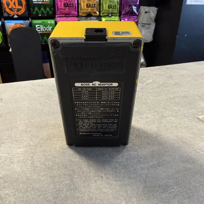 Pre-Owned Boss OD-1 Black and yellow power adapter on a gray surface with colorful boxes in the background